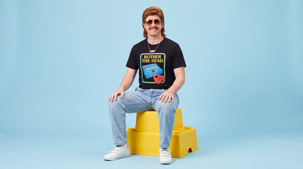 A studio portrait of a man with a mullet, moustache, and sunglasses sitting on a yellow plastic block against a pale blue background. He wears a gold chain, light blue jeans, and a t-shirt showing an illustration of a chunky, brightly colored plastic toy and a red planchette with bubbly letters reading 'BOTHER THE DEAD'. He is smiling warmly.